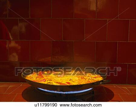 Stock Image - Steaming dish of paella cooking on gas stove. Fotosearch