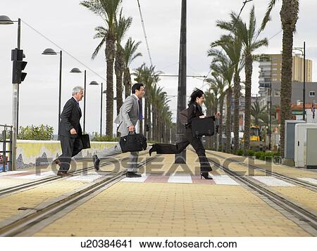 Stock Image - Businesswoman and two businessmen running acrossdouble tram lines with suitcases at zebra crossing. Alicante, Spain.. Fotosearch