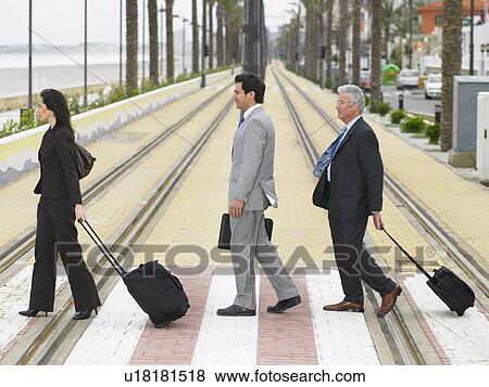 Stock Photo - Businesswoman and two businessmen crossing double tram lines with suitcases at zebra crossing. Alicante, Spain.. Fotosearch
