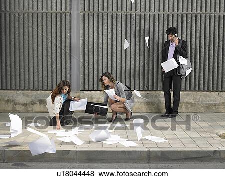 Two businesswomen picking up papers blowing about pavement while a young man looks on, talking on the phone, View Large Photo Image Stock Photo - Two businesswomen picking up papers blowing about pavement while a young man looks on, talking on the phone,. Fotosearch