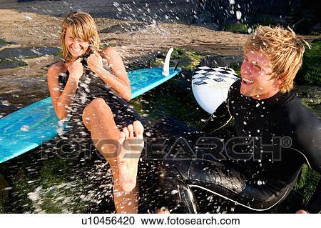 Stock Image - Couple splashing water on each other laughing with surfboards in background.. Fotosearch