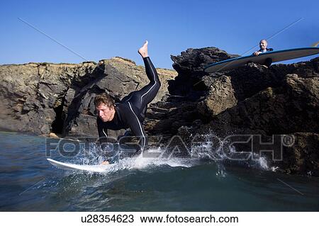 Man jumping onto surfboard with other man sitting on rocks behind him. View Large Photo Image Stock Image - Man jumping onto surfboard with other man sitting on rocks behind him.. Fotosearch