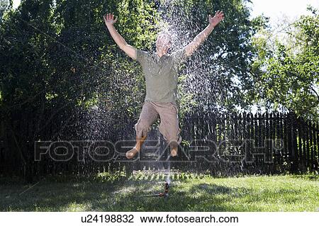 Stock Image - Man jumping through sprinkler.. Fotosearch