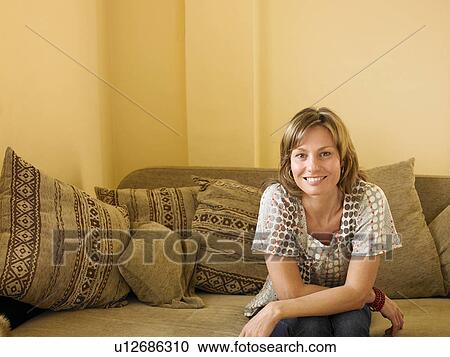 Stock Image - Woman sitting on sofa at home, smiling, front view, portrait. Fotosearch