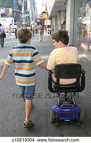 Picture - Brothers, one using a scooter for mobility, going down a city sidewalk.. Fotosearch