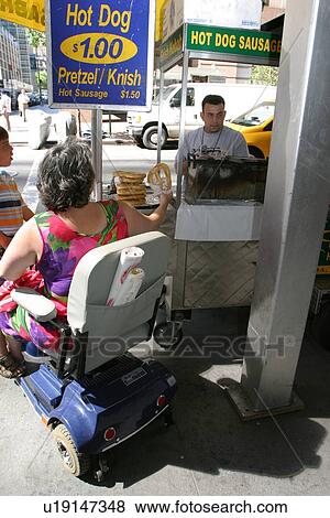 Stock Photo - Woman using a scooter/cart purchasing a hot dog from a street vendor in New York.. Fotosearch
