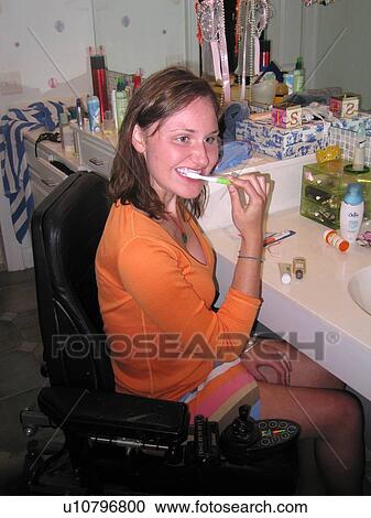 Stock Image - Young woman (electric wheelchair user) brushes her teeth in an accessible bathroom.. Fotosearch
