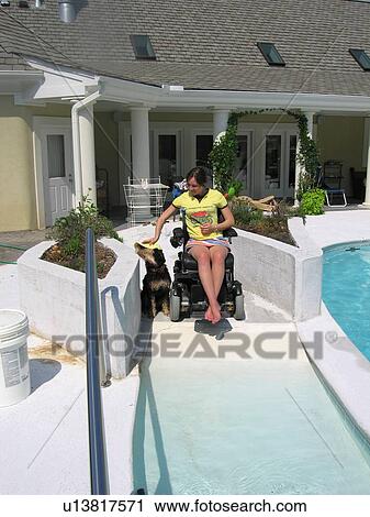 Stock Image - Young woman (electric wheelchair user) stops to pet dog at the top of the ramp entrance to her accessible pool.. Fotosearch