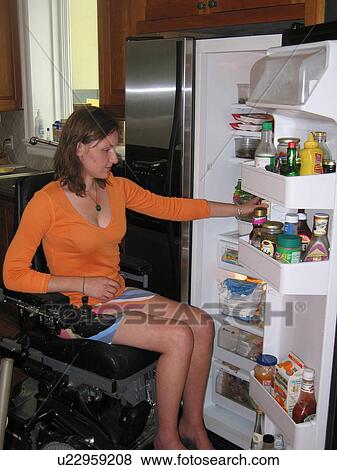 Stock Photo - Young woman (electric wheelchair user) selects items from an accessible refrigerator.. Fotosearch