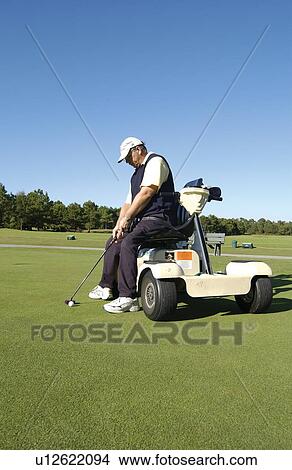 Picture - Man with a disability playing golf at a golf course while sitting on his scooter that he uses for mobility.. Fotosearch