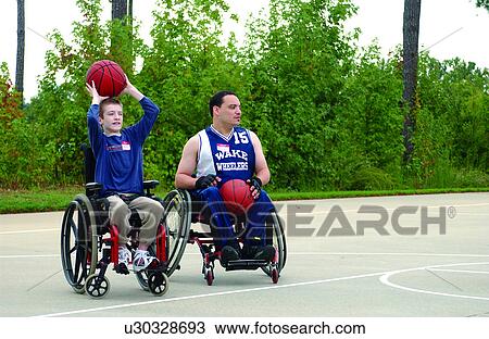 Stock Image - Two friends, both utilizing wheelchairs for mobility, playing a game of basketball at their local recreation center.. Fotosearch