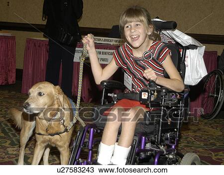 Stock Image - Smiling little girl with disabilities operating her electric wheelchair while holding the leash of her service dog.. Fotosearch