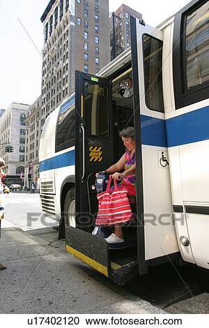 Stock Image - Woman with a disability (carrying a bright pink striped tote! ) exiting a city bus via a lift.. Fotosearch