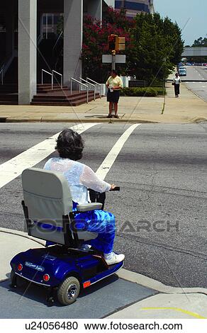 Stock Image - Woman with a disability using a motorized scooter for mobility crossing the street at a cross walk.. Fotosearch