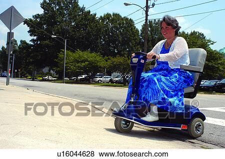 Stock Photo - Woman with a disability using a motorized scooter to navigate the streets in a city. Thankfully, this city has curb cuts which allows her access to the places she want to go to.. Fotosearch