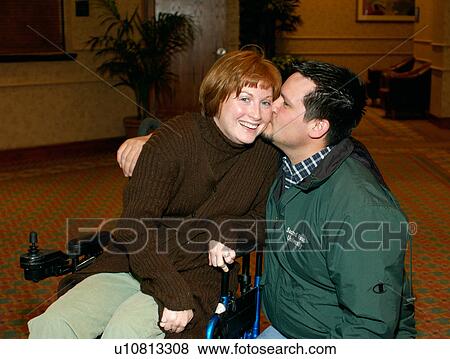 Stock Photo - Woman with a disability, utilizing a wheelchair for mobility, getting a kiss and hug from her husband.. Fotosearch