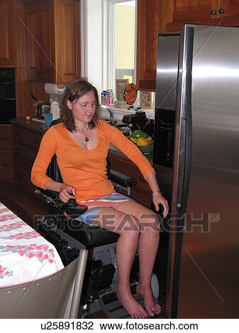 Stock Image - Young woman (electric wheelchair user) selects items from the refrigerator in an accessible kitchen.. Fotosearch