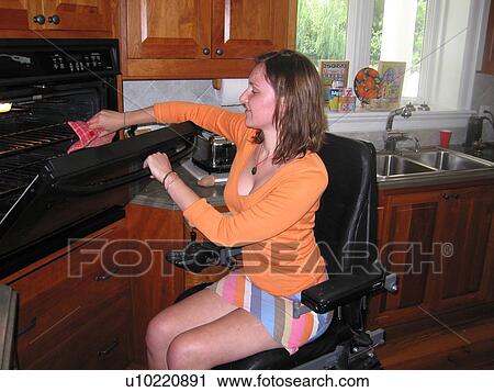 Stock Image - Young woman (wheelchair user) uses oven in her accessible kitchen.. Fotosearch
