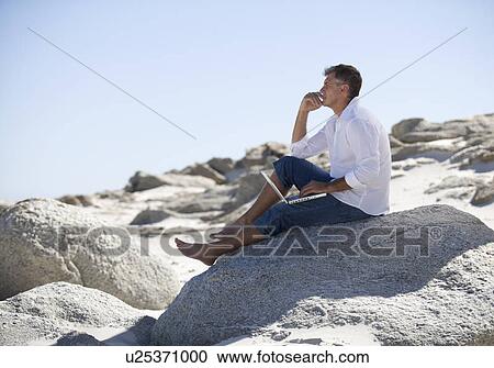 A man sitting on rocks working on a laptop View Large Photo Image Stock Image - A man sitting on rocks working on a laptop. Fotosearch