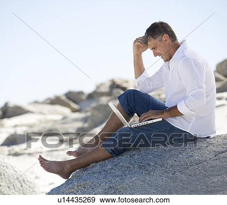 A man sitting on rocks working on a laptop View Large Photo Image Stock Photo - A man sitting on rocks working on a laptop. Fotosearch