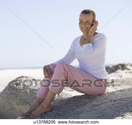 A mature woman sitting on a beach using a mobile phone View Large Photo Image Stock Photograph - A mature woman sitting on a beach using a mobile phone. Fotosearch