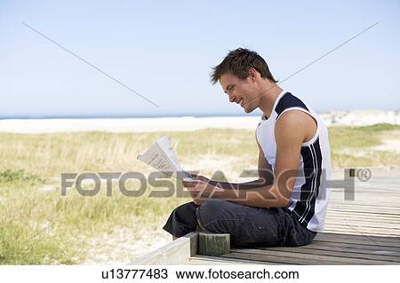 Stock Image - A young man sitting on a beach reading a newspaper. Fotosearch