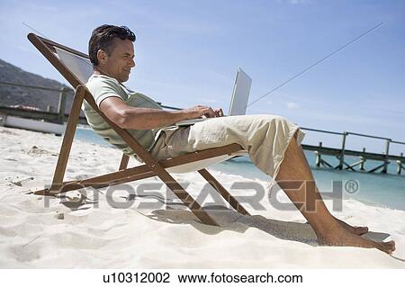 Stock Image - A mature man sitting in a deck chair using a laptop. Fotosearch