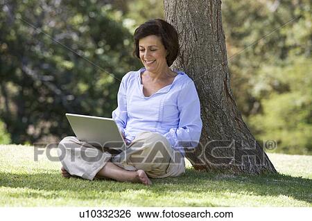 A senior woman sitting under a tree using a laptop View Large Photo Image Stock Photograph - A senior woman sitting under a tree using a laptop. Fotosearch