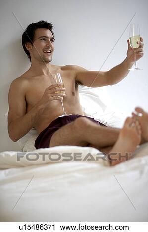 Stock Image - Young man holding two champagne glasses, sitting on a bed. Fotosearch