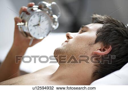 Young man looking at his alarm clock View Large Photo Image Stock Image - Young man looking at his alarm clock. Fotosearch