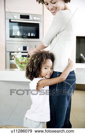Stock Photo - Mother making salad lunch in the kitchen while young daughter gives her a hug. Fotosearch