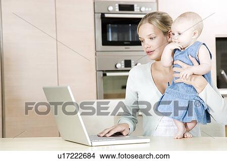 Mother using a laptop in the kitchen at home, holding baby daughter View Large Photo Image Picture - Mother using a laptop in the kitchen at home, holding baby daughter. Fotosearch