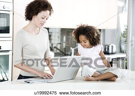 Mother using a laptop in the kitchen at home, while young daughter looks on View Large Photo Image Stock Photography - Mother using a laptop in the kitchen at home, while young daughter looks on. Fotosearch