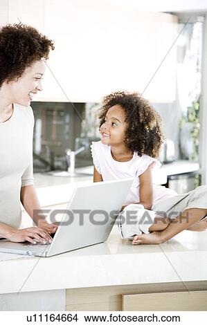 Mother using laptop in kitchen while young daughter looks on smiling View Large Photo Image Picture - Mother using laptop in kitchen while young daughter looks on smiling. Fotosearch