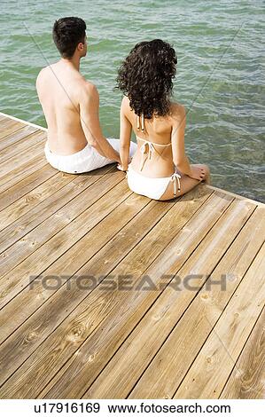 Couple sitting on the side of a wooden jetty by the sea, holding hands View Large Photo Image Stock Photo - Couple sitting on the side of a wooden jetty by the sea, holding hands. Fotosearch