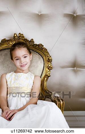 Stock Image - Little girl sitting in an ornate chair wearing a party dress. Fotosearch