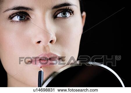 Stock Image - Young woman putting on lipstick in front of a mirror. Fotosearch