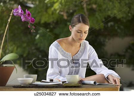 Stock Photo - A young woman sitting at a table reading a magazine. Fotosearch