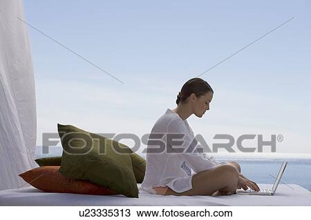 A young woman sitting on a bed with a laptop View Large Photo Image Stock Image - A young woman sitting on a bed with a laptop. Fotosearch