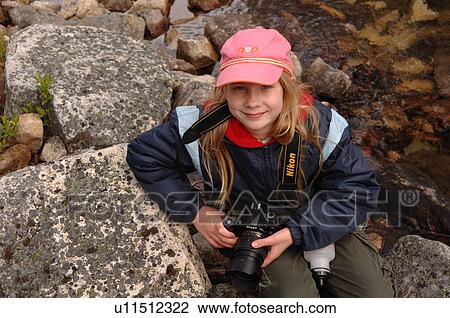 Stock Image - Girl holding camera sitting amongst rocks. Fotosearch