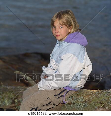 Stock Image - Side profile of a young girl sitting on a fallen tree looking at camera. Fotosearch
