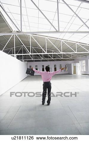 Stock Photograph - Rear view of a businessman standing with his arms outstretched and holding a briefcase in an entrance hall. Fotosearch