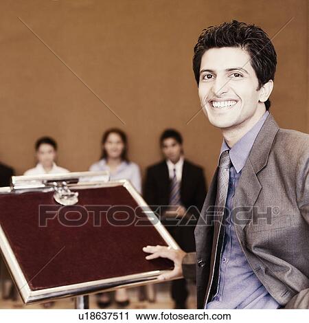 Businessman standing at a lectern with his colleagues in the background View Large Photo Image Stock Image - Businessman standing at a lectern with his colleagues in the background. Fotosearch
