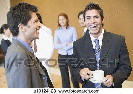 Close-up of two businessmen smiling with their colleagues standing behind them View Large Photo Image Stock Image - Close-up of two businessmen smiling with their colleagues standing behind them. Fotosearch