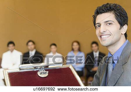 Stock Image - Portrait of a businessman smiling at a lectern with his colleagues in the background. Fotosearch