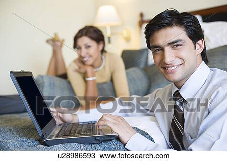 Portrait of a businessman working on a laptop and with a young woman lying on the bed in the background View Large Photo Image Stock Image - Portrait of a businessman working on a laptop and with a young woman lying on the bed in the background. Fotosearch