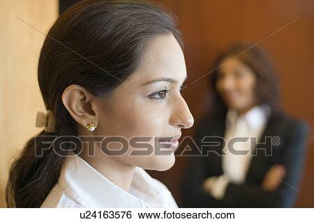 Stock Photograph - Side profile of a businesswoman smiling with another businesswoman standing in the background. Fotosearch