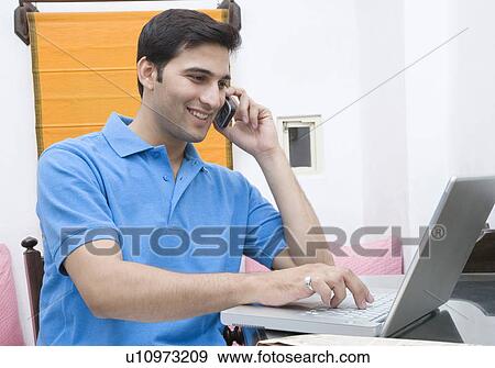 Close-up of a young man talking on a mobile phone and using a laptop View Large Photo Image Stock Photo - Close-up of a young man talking on a mobile phone and using a laptop. Fotosearch