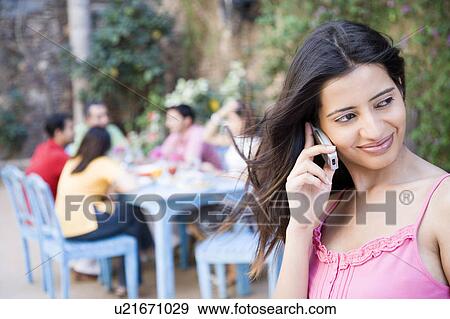 Close-up of a young woman talking on a mobile phone with her friends sitting at a dining table in the background View Large Photo Image Stock Photo - Close-up of a young woman talking on a mobile phone with her friends sitting at a dining table in the background. Fotosearch