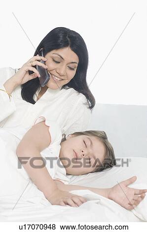 Stock Photo - Girl sleeping on the bed with her mother talking on a mobile phone beside her. Fotosearch
